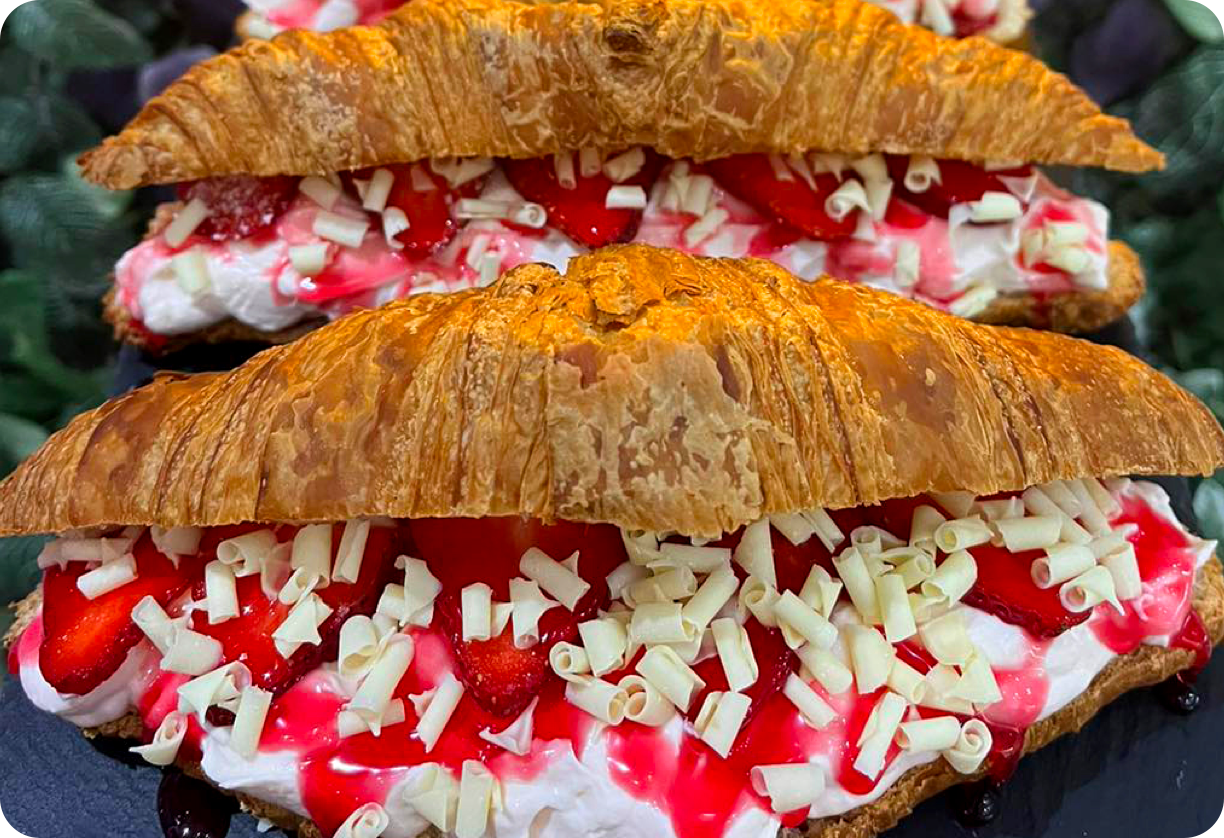 Image is of three croissants, each filled with cheesecake filling, fresh strawberries, strawberry sauce and white chocolate curls. A neon sign in the shape of an ice cream is just visible in the background.